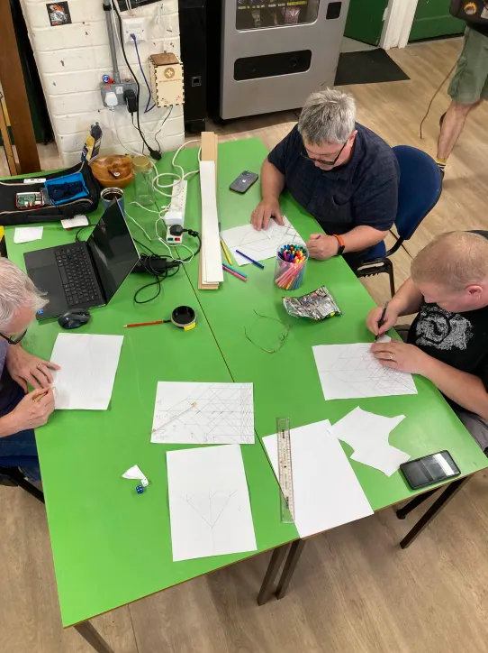 A photo of 3 people sat at a green table. They hold pencils and look intently at their work, plotting lines on pieces of paper. The photo is taken from a high position, and you can see completed phase graphs, laptops and other bits and bobs on the table.