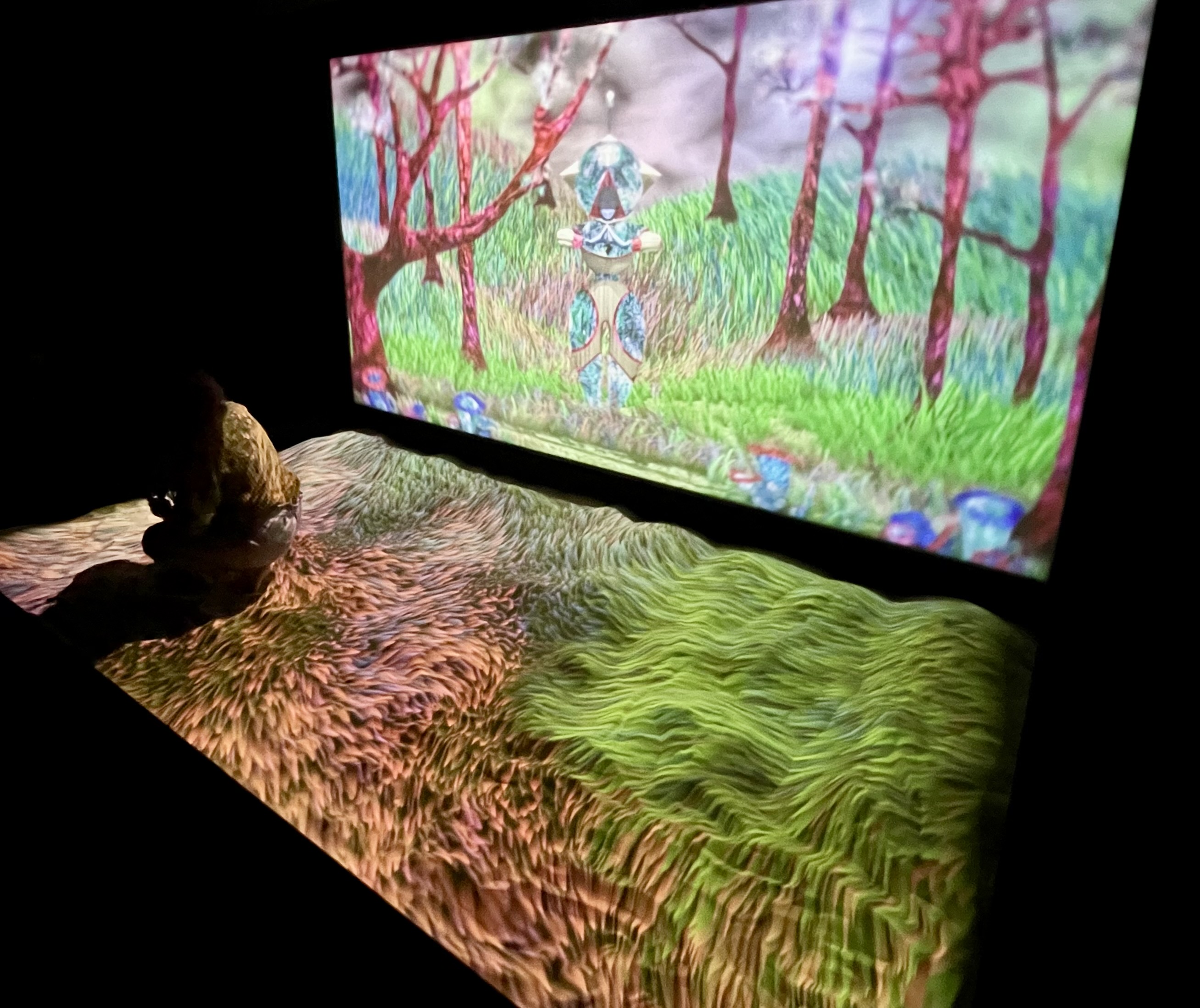 A person crouches in a massive sandpit that has thousands of blades of grass projected onto it waving in different colours. The wall behind shows a matching video of an unreal grassy hillside with trees and a standing figure.