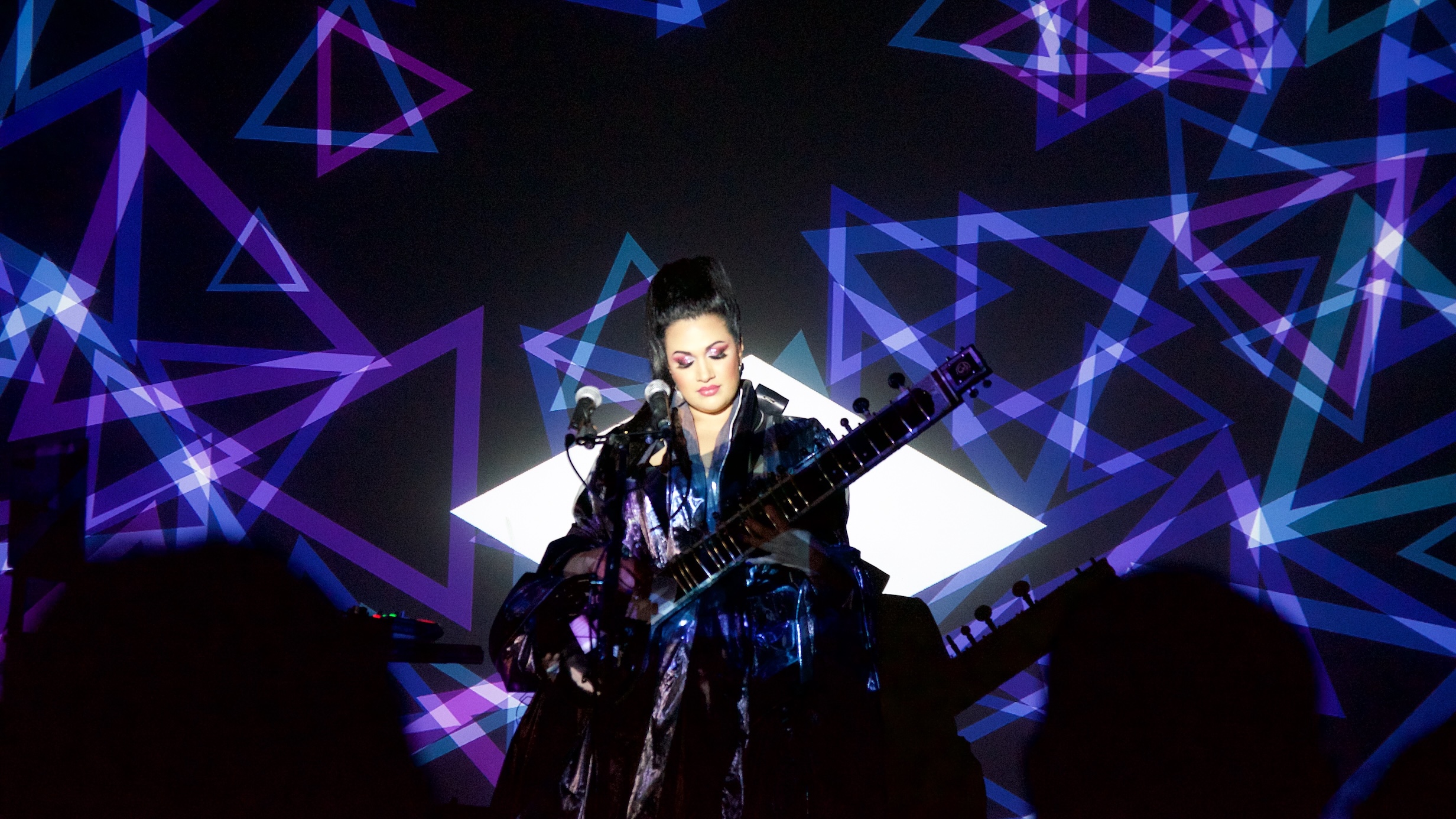 The artist BISHI, her face illuminated by a white diamond-shaped spotlight, plays a sitar in front of a projected screen of dozens of large blue and purple outlined triangles